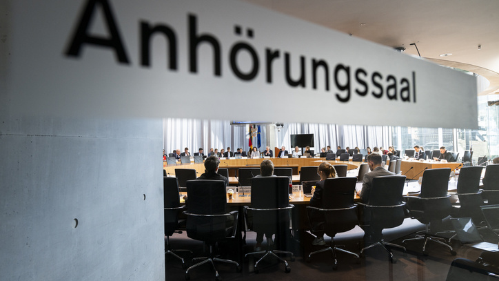 Aufschrift Anhörungssaal mit Blick in den Saal, wo ein Kreis von Politiker*innen sitzt