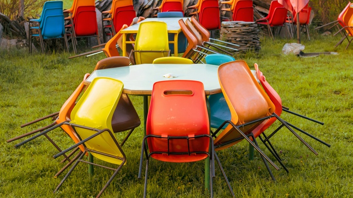 Ein Kreis aus bunten Stühlen lehnt auf einer Wiese an einem Tisch.