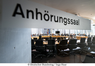Aufschrift Anhörungssaal mit Blick in den Saal, wo ein Kreis von Politiker*innen sitzt