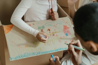 Zwei personen malen mit Buntestiften ein Bild von einem Haus.
